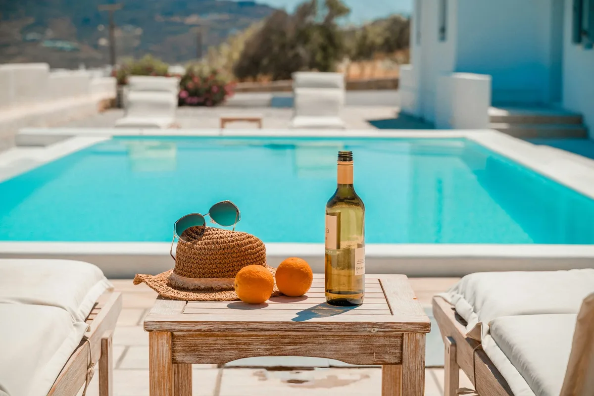 A poolside table in the sun: a straw hat, sunglasses, two oranges and a bottle of wine, with an empty turquoise pool behind.