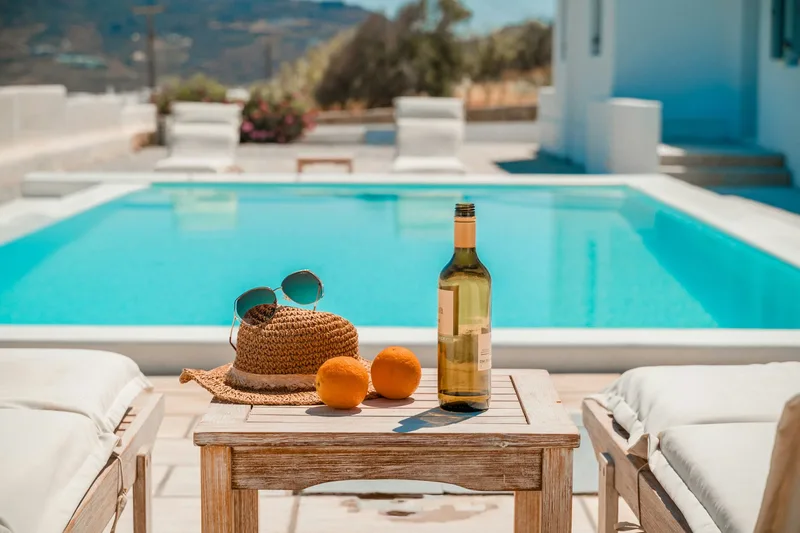 A poolside table in the sun: a straw hat, sunglasses, two oranges and a bottle of wine, with an empty turquoise pool behind.