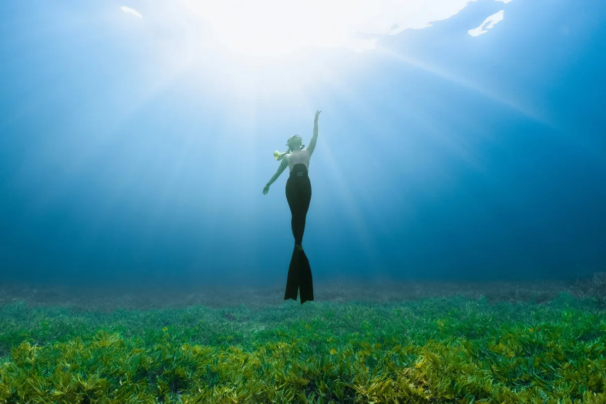 A freediver hangs weightless in clear blue water above a meadow of seagrass, sunbeams fanning down from the surface.