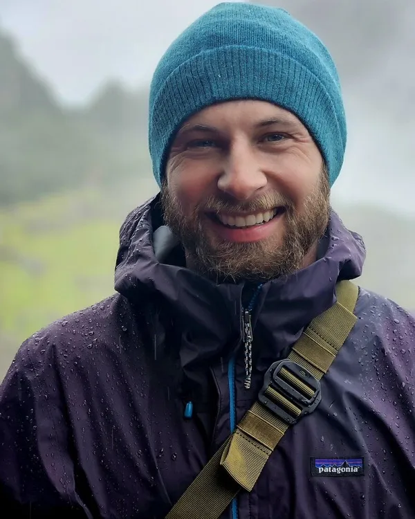 Eric, smiling in a teal beanie and rain jacket on a misty green hillside.